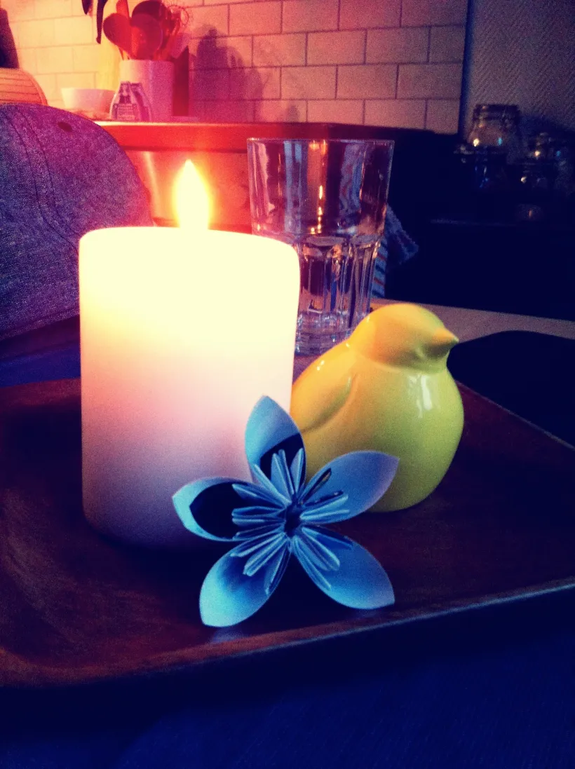 Tent lys, et glass, en keramisk fuglefigur og en blå origamiblomst på en trebrett, varm og rolig stemning