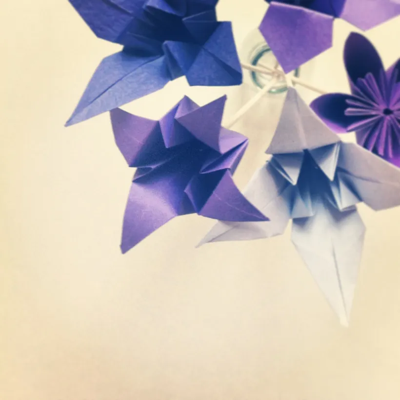 Lilla og blå origamiblomster mot en myk kremfarget bakgrunn, dempede farger og en sart håndverksstemning