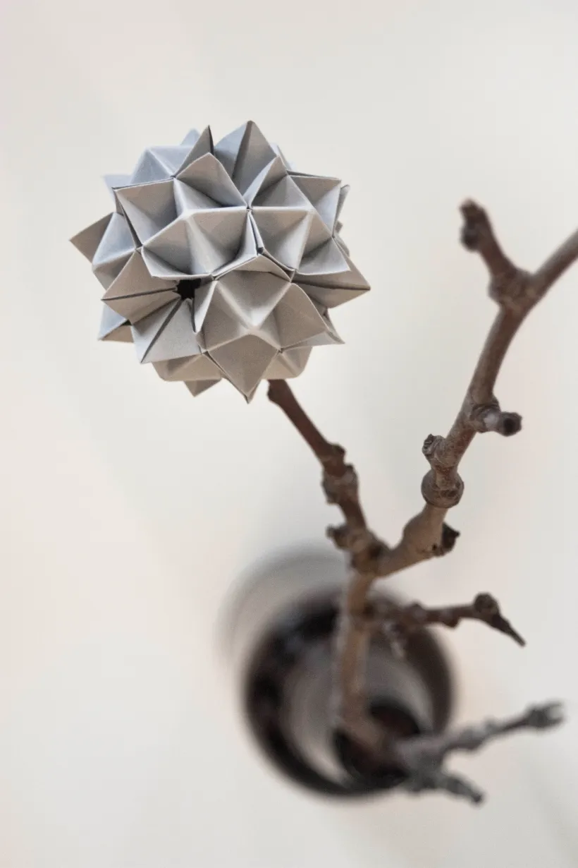 Nærbilde av en grå geometrisk papirskulptur formet som en spiss blomst, festet til en bar gren i en mørk vase